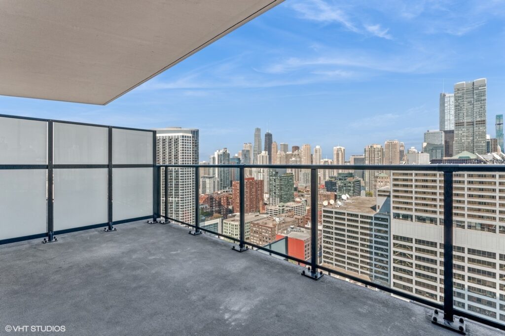 High-rise building balcony with glass railing, offering a view of a cityscape with skyscrapers under a clear blue sky.