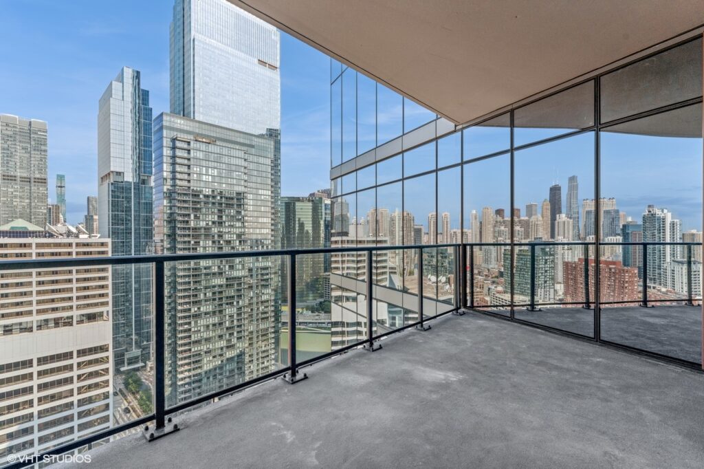 Spacious balcony with glass railing overlooking a cityscape featuring modern skyscrapers and a clear blue sky.