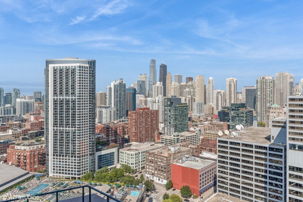 City skyline with numerous skyscrapers under a blue sky. A mix of modern and older buildings, with various heights and designs, are visible, along with some greenery and light traffic below.