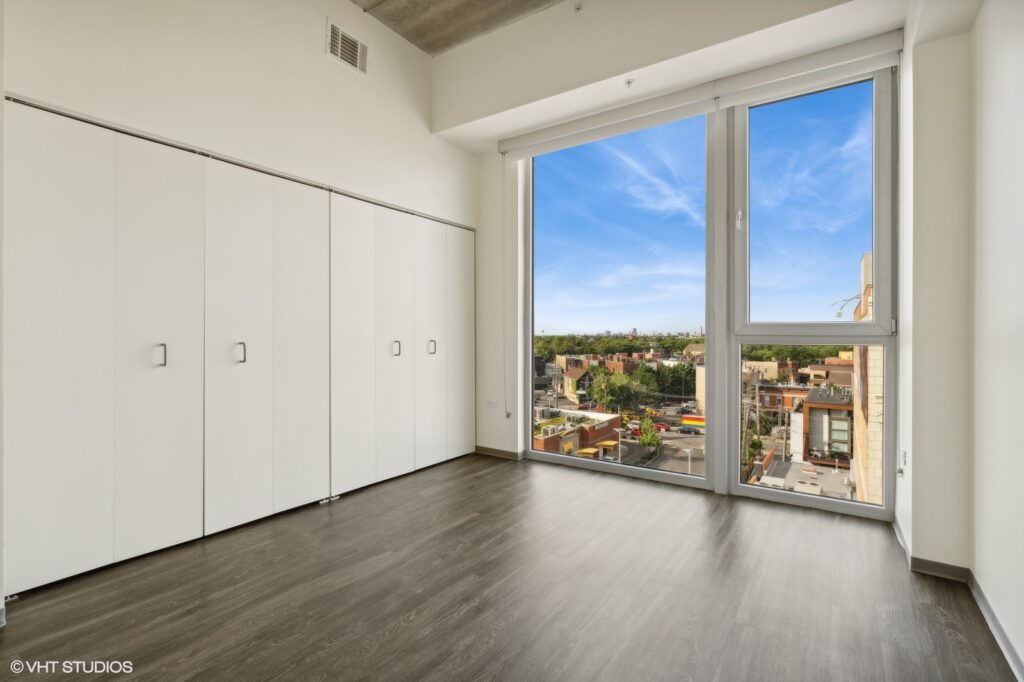 A bright, empty room with a wall of white closets and a large window overlooking a cityscape under a blue sky. Dark wood flooring is present.