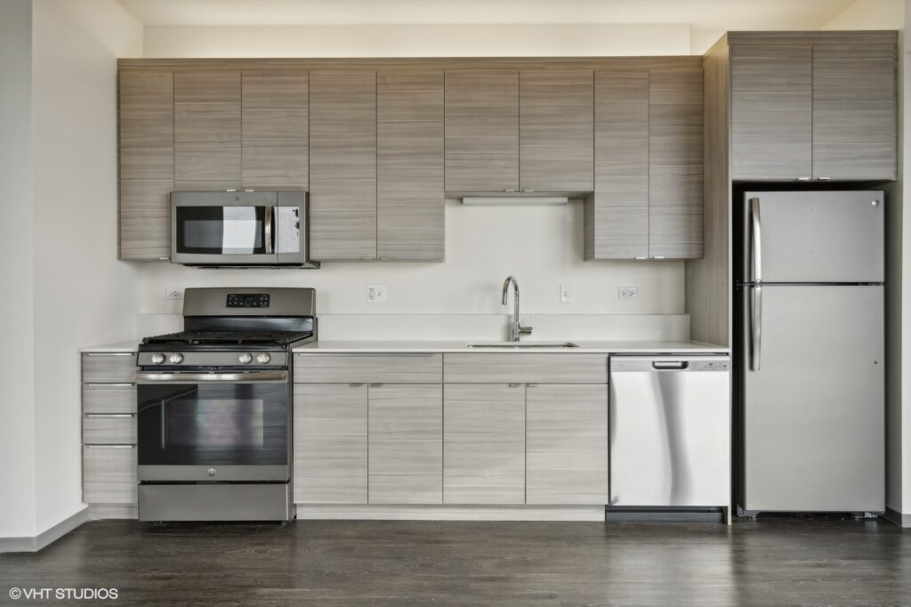 Modern kitchen with light wood cabinets, stainless steel appliances, including a microwave, oven, dishwasher, and fridge, and a white backsplash.