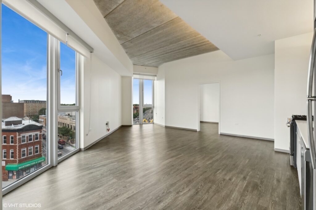Spacious empty apartment with large windows, wooden floors, and a view of a city street. Ceiling has a modern concrete finish. Kitchen area with stainless steel appliances visible on the right.