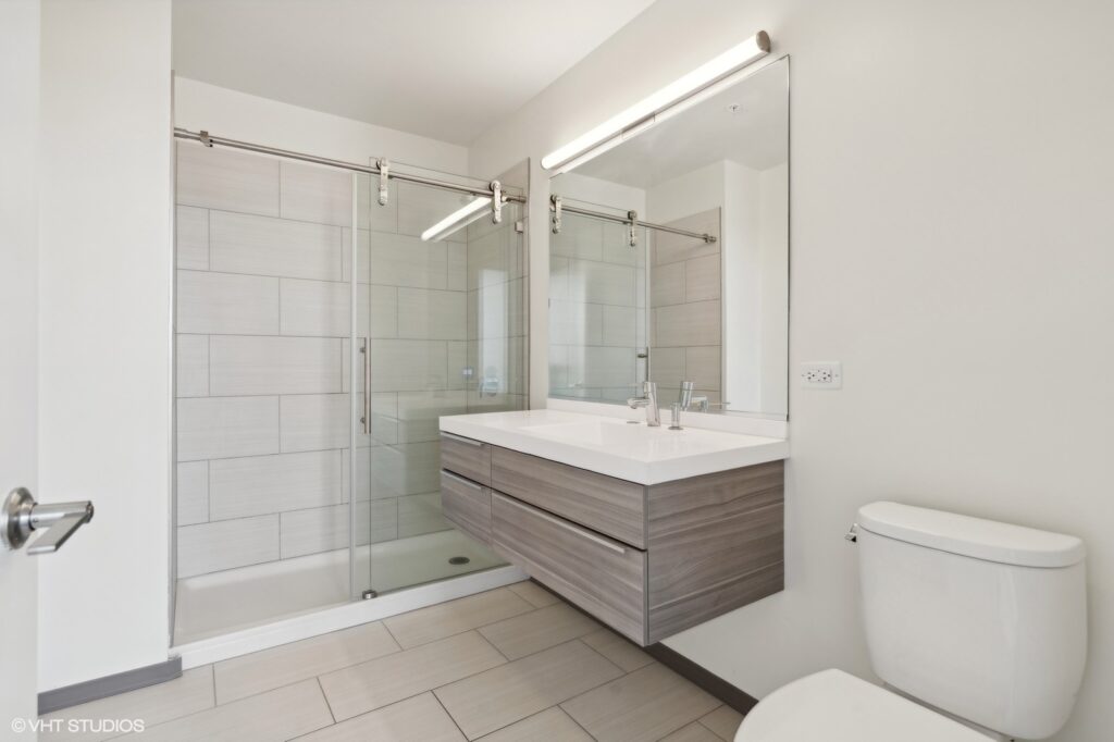 Modern bathroom with a glass-enclosed shower, floating vanity with dual sinks, large mirror, and a toilet. Walls and floor are tiled in neutral tones.