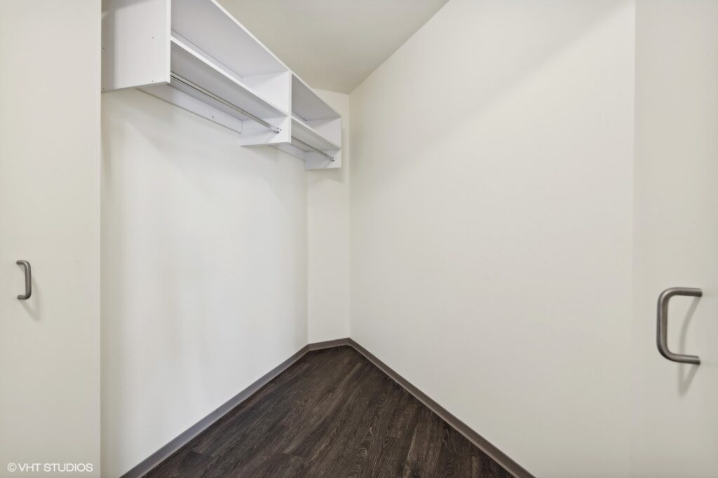 Empty walk-in closet with white walls, dark wood flooring, and white shelving on the left wall.