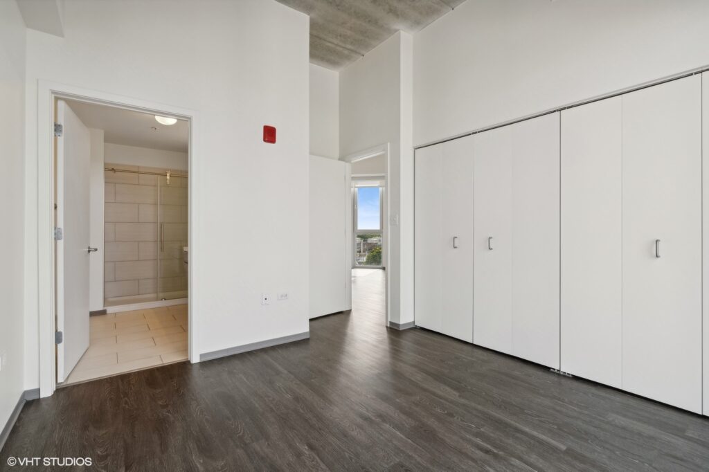 Empty room with dark wood flooring, a white closet, and a bathroom with a tiled shower visible through an open door. Ceiling is concrete and walls are white. Light enters through a distant window.