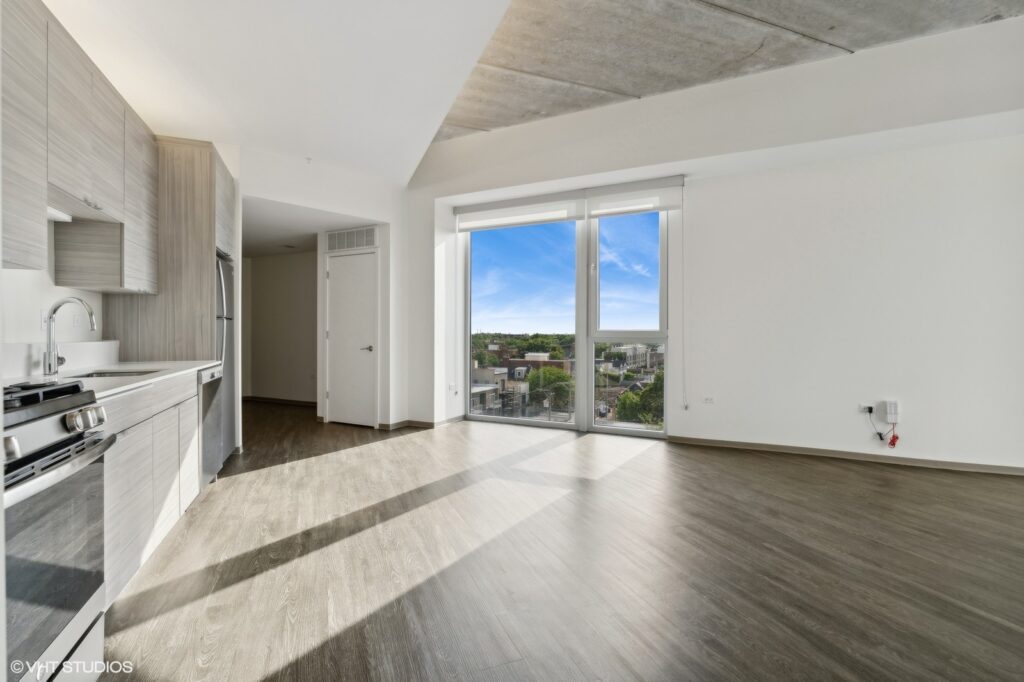 Modern, unfurnished studio apartment with wood floors, large windows, and view of trees and sky. Contains kitchen with stainless steel appliances on the left side.