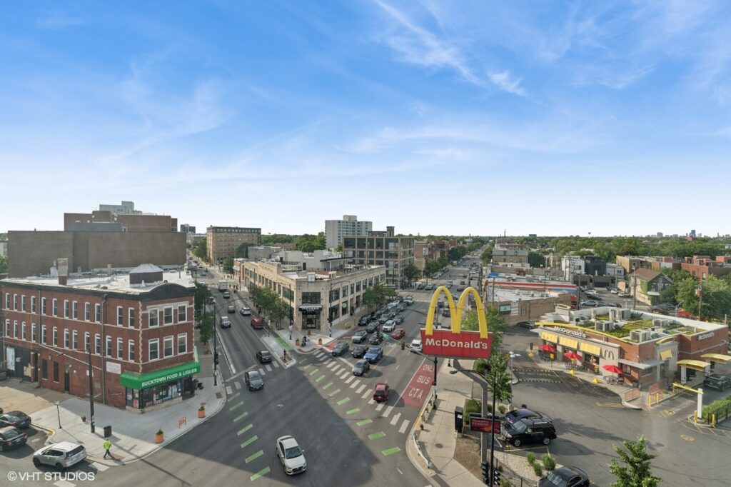 Aerial view of a busy urban intersection with multiple cars. A McDonald's restaurant with its distinctive yellow arches is prominently visible. Rows of buildings line the streets under a clear blue sky.