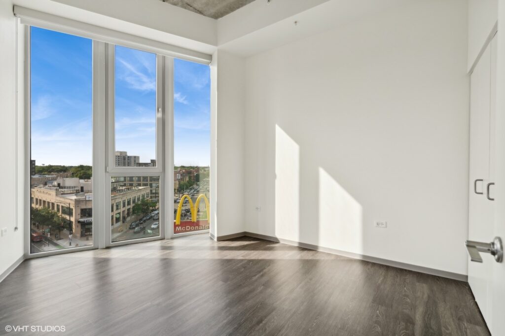 Bright, empty room with large windows showing a cityscape view including a McDonald's sign. Wood floor and white walls are visible.