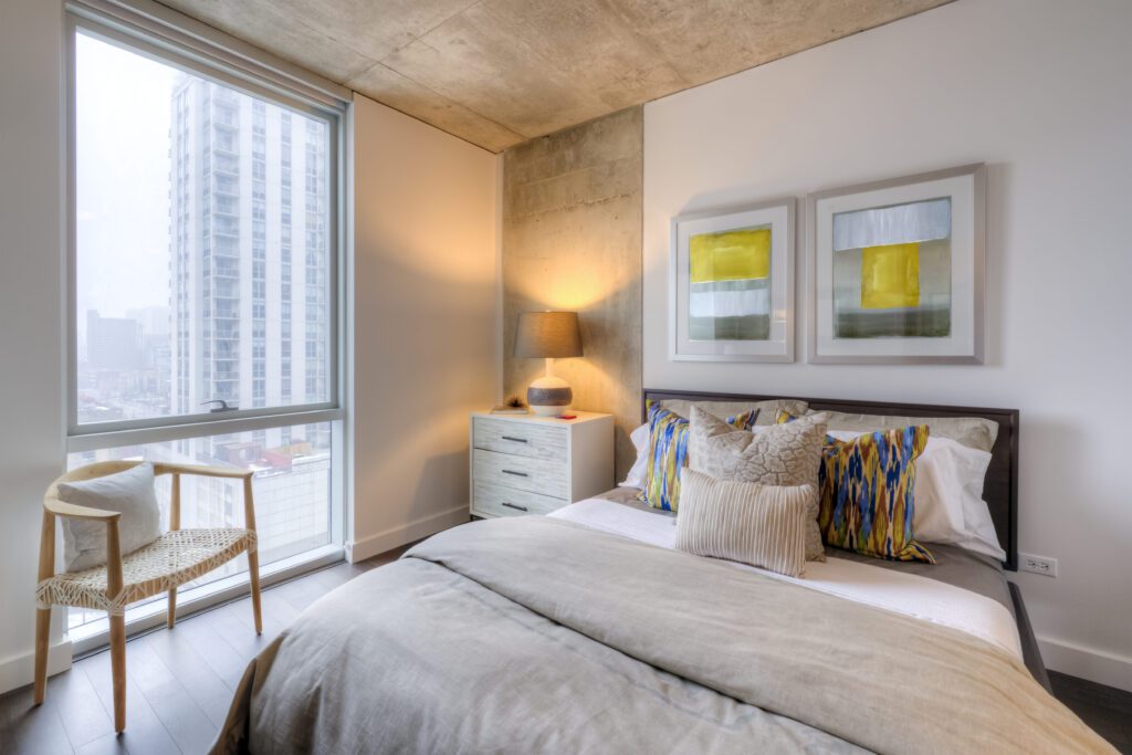 A modern bedroom with a large window, showing a tall building outside. Features a double bed with colorful pillows, wall art, a bedside lamp, a chair, and a wooden ceiling.