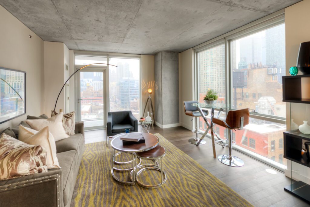 Modern living room with large windows, city view, and contemporary furnishings. Includes a sofa, armchair, coffee table, and bar stools at a small table. Neutral tones and bright light.