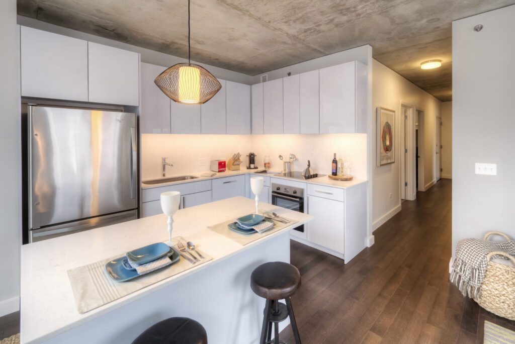Modern kitchen with white cabinets, stainless steel appliances, and pendant light. Two place settings on the island with stools and a basket on wooden floor in the background.