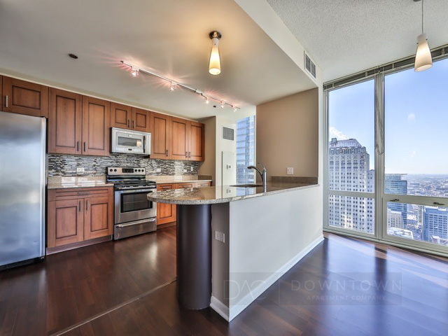 Modern kitchen with wooden cabinets, stainless steel appliances, granite countertops, and dark wood flooring. Large windows offer a city view. Ceiling lights provide illumination.