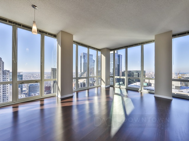 Spacious corner apartment with floor-to-ceiling windows, wooden floors, and city skyline views in a modern high-rise building.