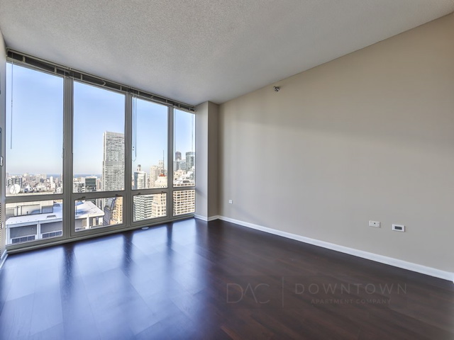 An empty apartment with large floor-to-ceiling windows, offering a city view. The room has dark wood flooring and beige walls.