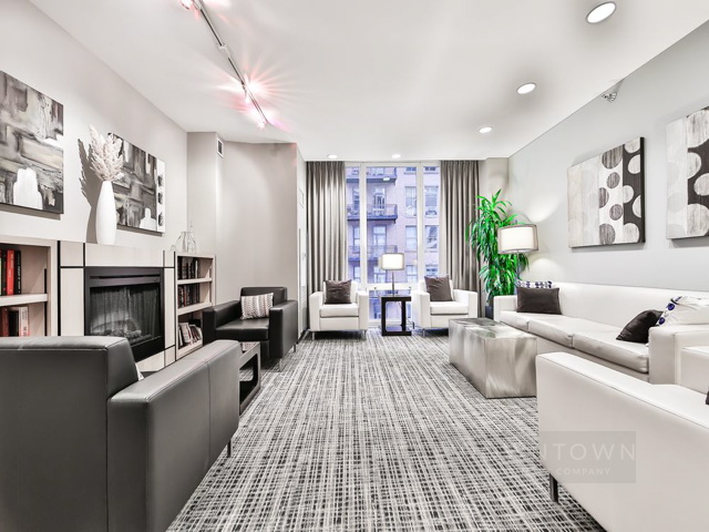 Modern living room with white and black furniture, abstract wall art, and a large window with city view. Patterned carpet and recessed lighting create a contemporary atmosphere.
