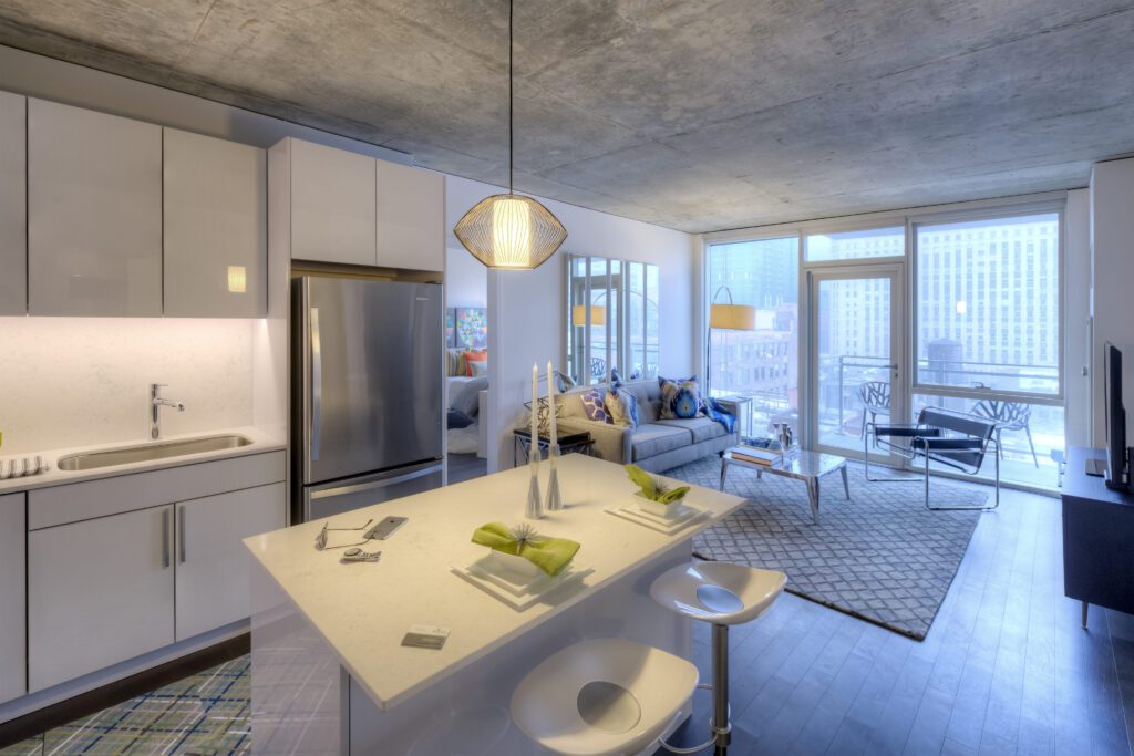 Modern open-concept apartment with kitchen island, bar stools, stainless steel appliances, and living area featuring a couch, coffee table, and large windows overlooking city buildings.