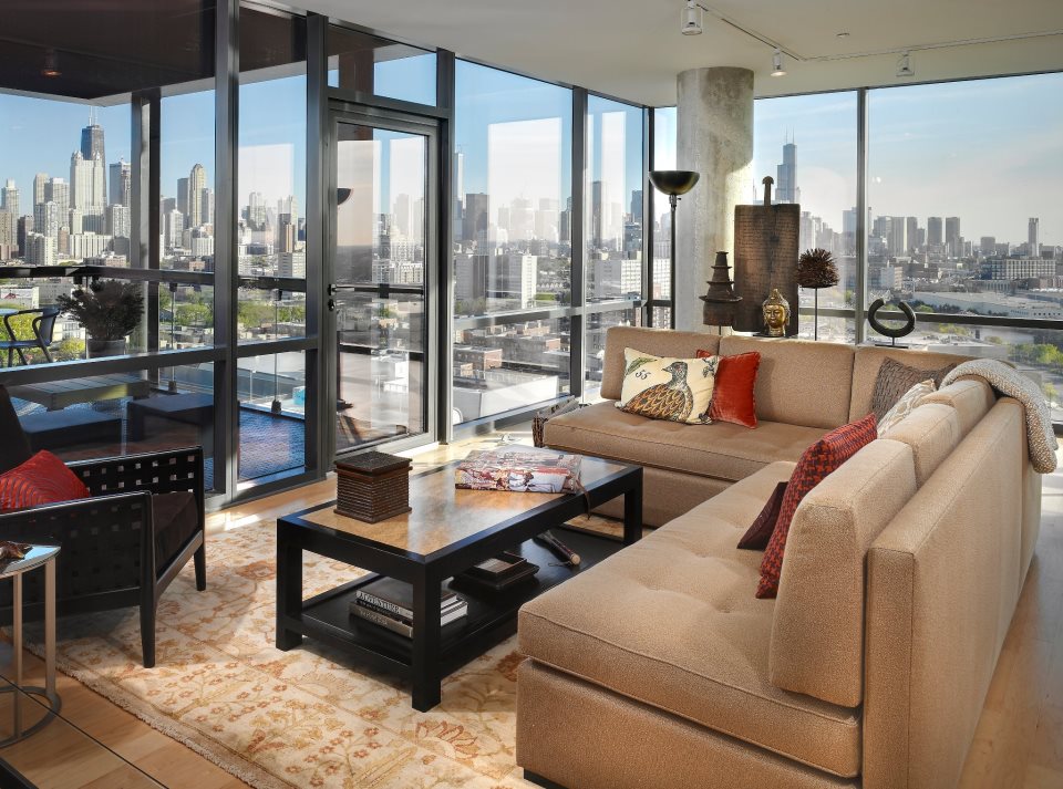 Modern living room with large beige sectional sofa, geometric coffee table, and floor-to-ceiling windows overlooking a cityscape. Neutral tones and eclectic decor elements enhance the space.