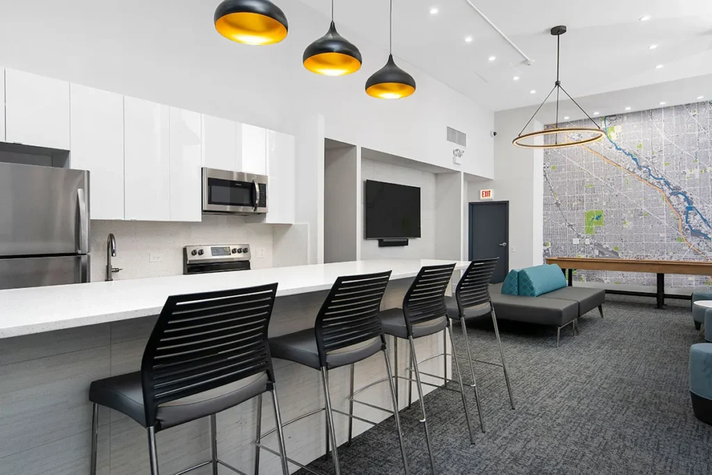 Modern kitchenette with bar seating and pendant lights. Features a stainless steel fridge, wall-mounted TV, and map artwork. Black chairs line the white counter. Gray couches in the lounge area.