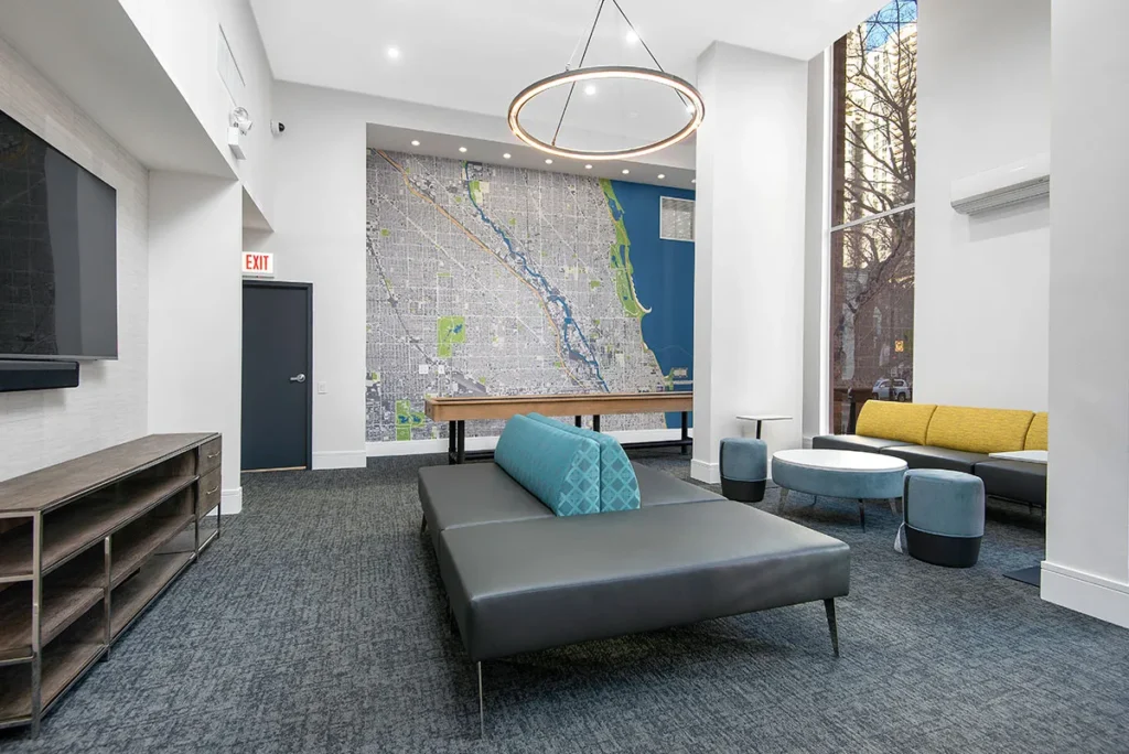 Modern lobby with gray seating, a wall map, large window, and overhead chandelier. Includes a TV on a stand, a table, and round chairs. Carpeted floor and emergency exit visible.