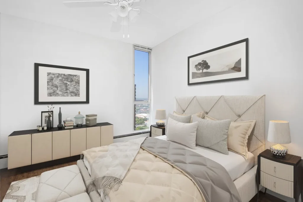 A modern bedroom with a large bed, beige bedding, two nightstands with lamps, a dresser with decor, and two framed pictures. A window offers a city view.