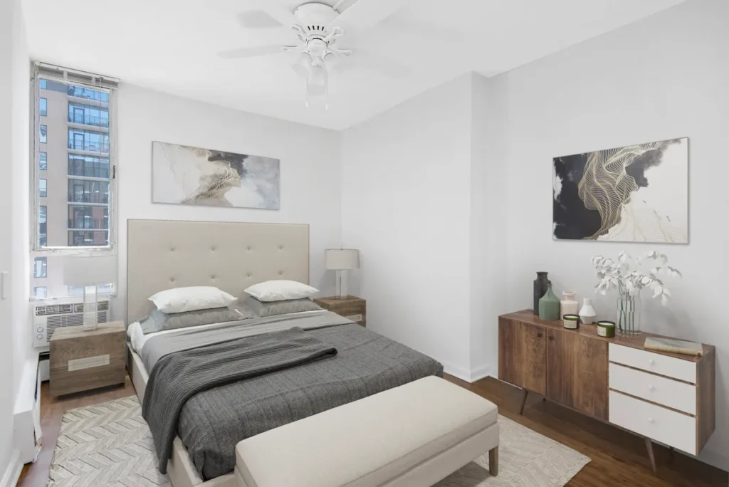 Modern bedroom with a double bed, beige headboard, and gray bedding. Wooden dresser and nightstand with decorative items. Two abstract paintings on the wall and a ceiling fan above.