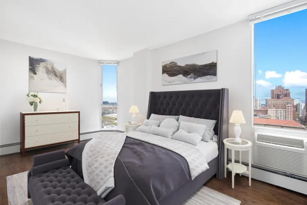 Bright modern bedroom with a large bed, gray upholstery, two side tables with lamps, a dresser, artwork on the walls, and city views from multiple windows.