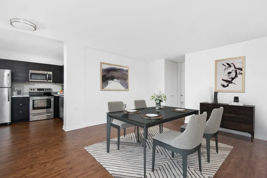 A modern dining room with a black table set for four on a striped rug. There's a dark wooden sideboard with art above it, and a kitchen with stainless steel appliances in the background.