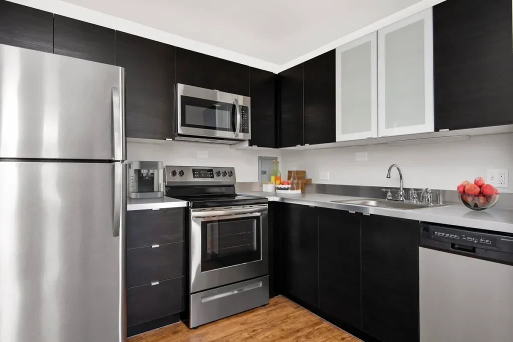 Modern kitchen with stainless steel appliances, dark cabinetry, and wooden flooring. Features a refrigerator, microwave, oven, dishwasher, and a sink. A bowl of apples is on the counter.
