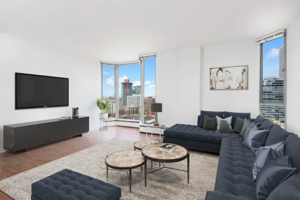 Modern living room with large sectional sofa, two round marble-topped tables, a wall-mounted TV, and city views through corner windows. Walls are white, and floor is hardwood.