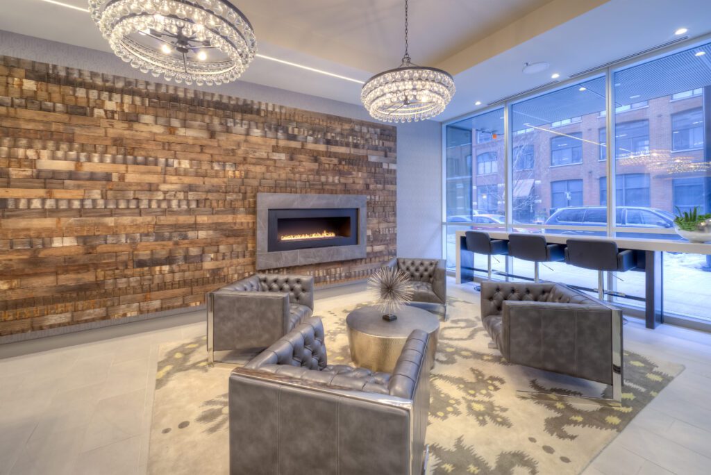 Modern lounge with four gray chairs around a round table, a wall-mounted fireplace on a wooden panel wall, large windows, and two decorative chandeliers.
