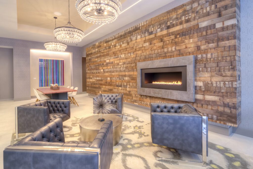 Modern lounge with a brick accent wall, linear fireplace, round wooden table, four gray quilted chairs, abstract wall art, and two large chandeliers.