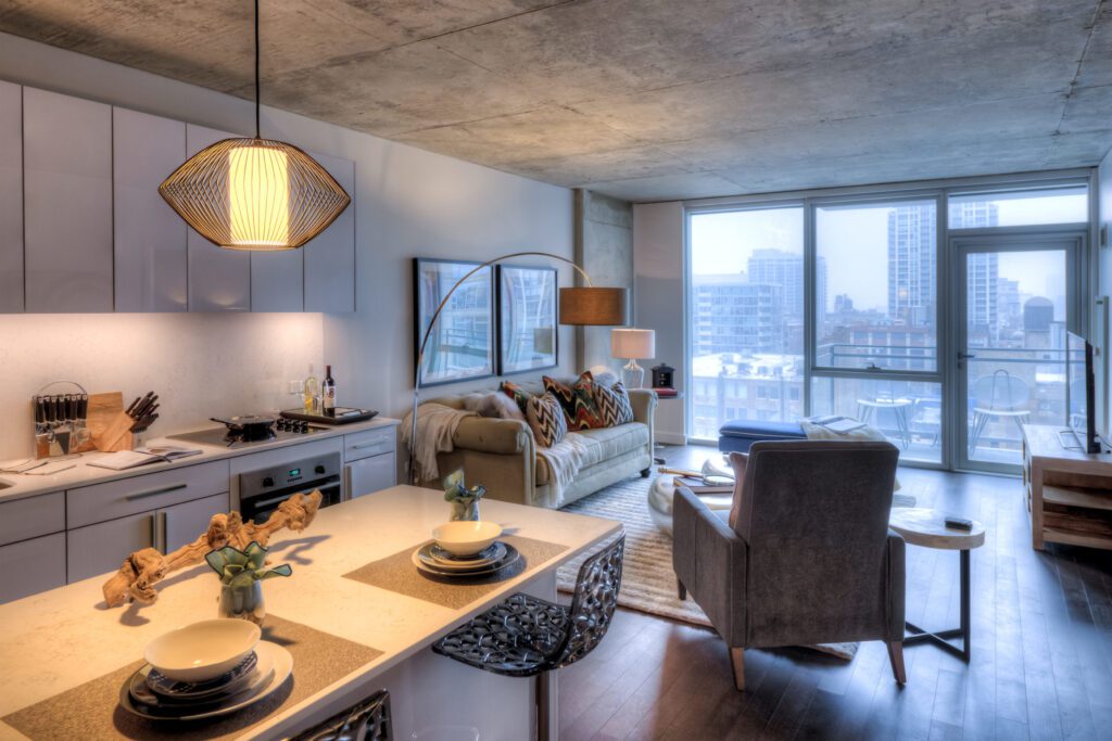 Modern apartment interior featuring a kitchen with an island, a living area with a sofa and armchair, large windows with city views, and contemporary decor.