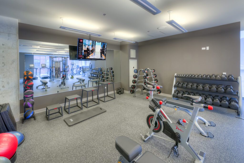 Modern gym with exercise bikes, dumbbells, medicine balls, and a wall mirror. A TV is mounted above the mirror. The room has gray walls, overhead lighting, and a carpeted floor.