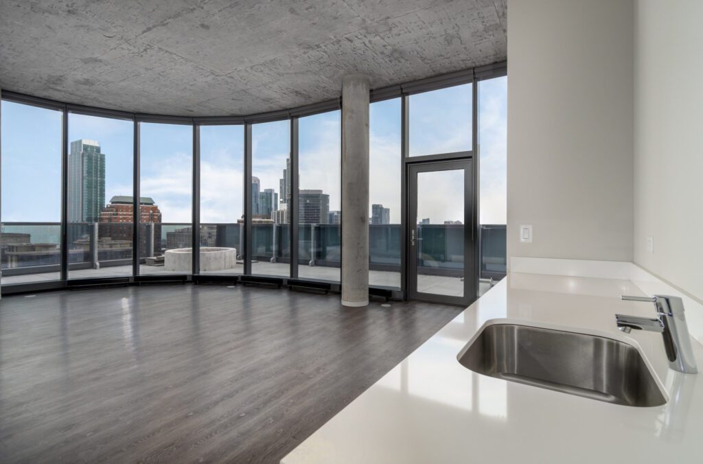 A modern, empty apartment with floor-to-ceiling windows showing a cityscape. The room features a concrete ceiling, dark wood flooring, and a white countertop with a sink.