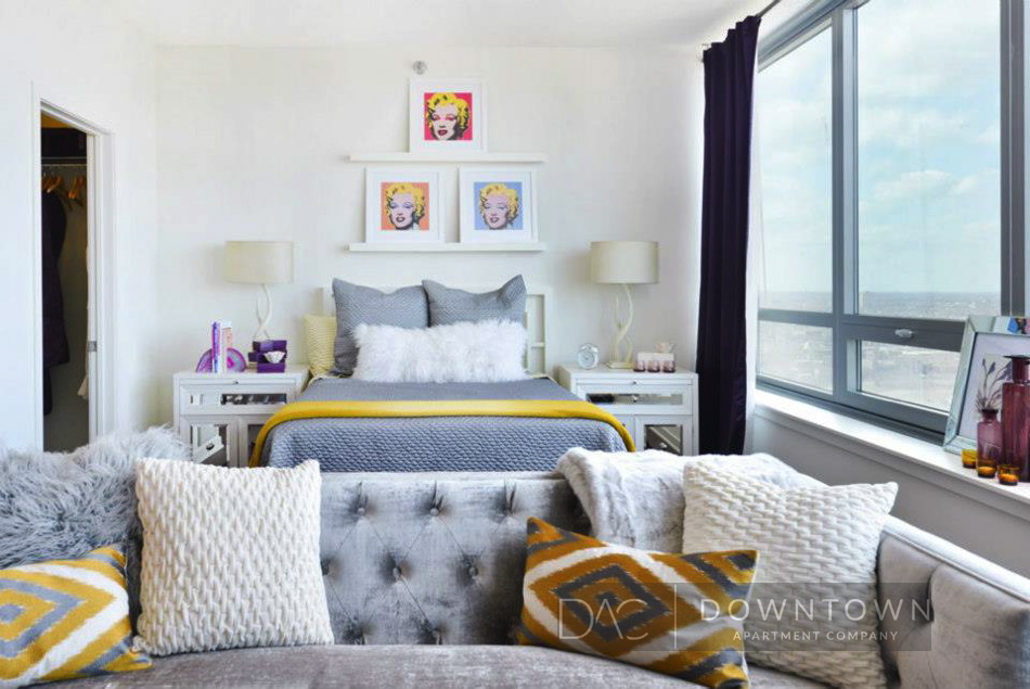 Stylish bedroom with a bed, decorative pillows, and pop art. Large window on the right wall. Shelves above the bed display art. Neutral tones with yellow accents.