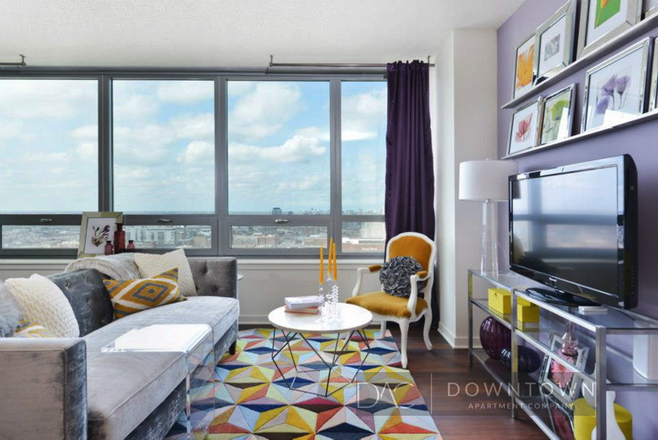Modern living room with large windows, gray sofa, geometric rug, TV, and decorative items. Purple curtains and a gallery wall with colorful art enhance the space.