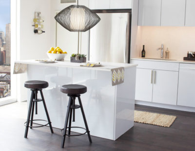 Modern kitchen with a white island, two bar stools, pendant light, and a bowl of lemons. Large window offers a city view.