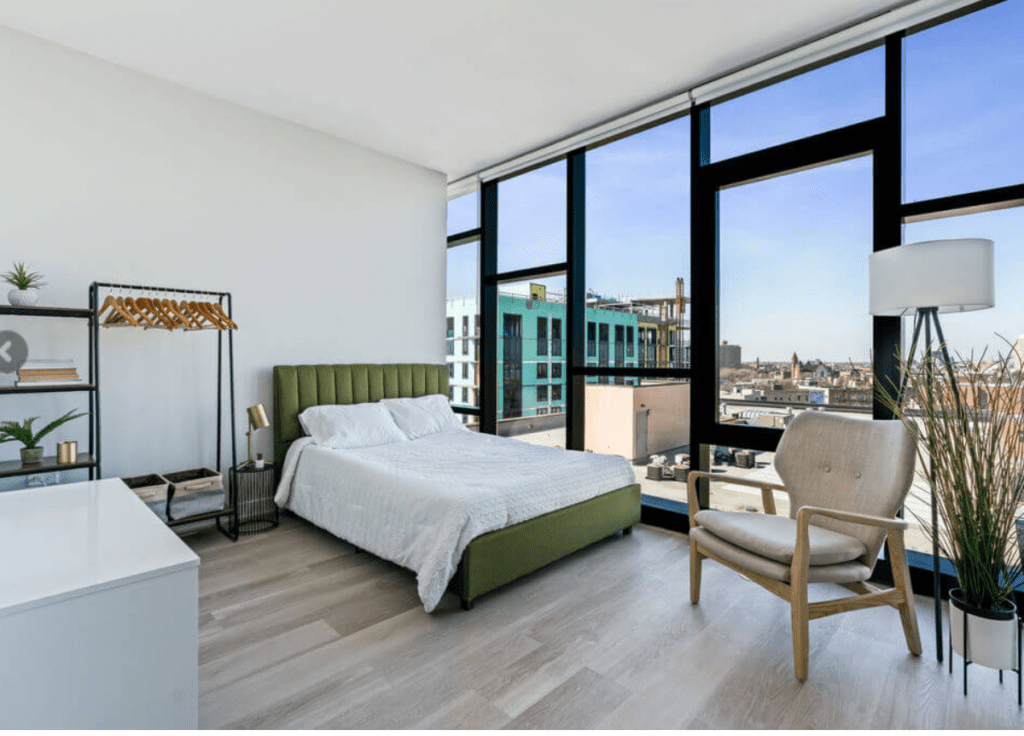 Modern bedroom with large windows, green upholstered bed, white bedding, minimalist decor, a floor lamp, and a wooden chair overlooking an urban view.