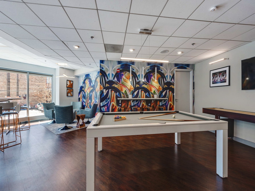 A recreation room featuring a pool table, vibrant mural artwork, seating area with blue chairs, tall windows, and a shuffleboard table on wooden flooring.