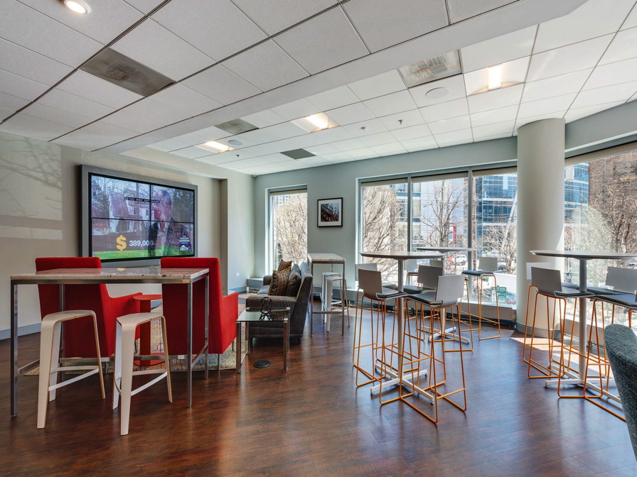 Modern lounge area with tall and low seating, tables, large windows, a wall-mounted TV, and hardwood floors, illuminated by natural light.
