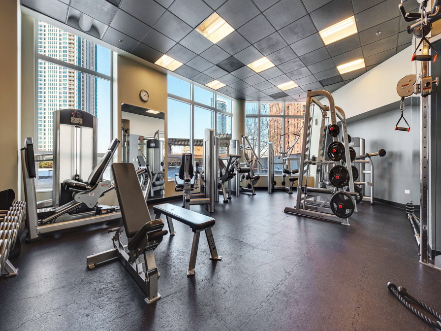 A gym with large windows features various weightlifting equipment, benches, and mirrors.
