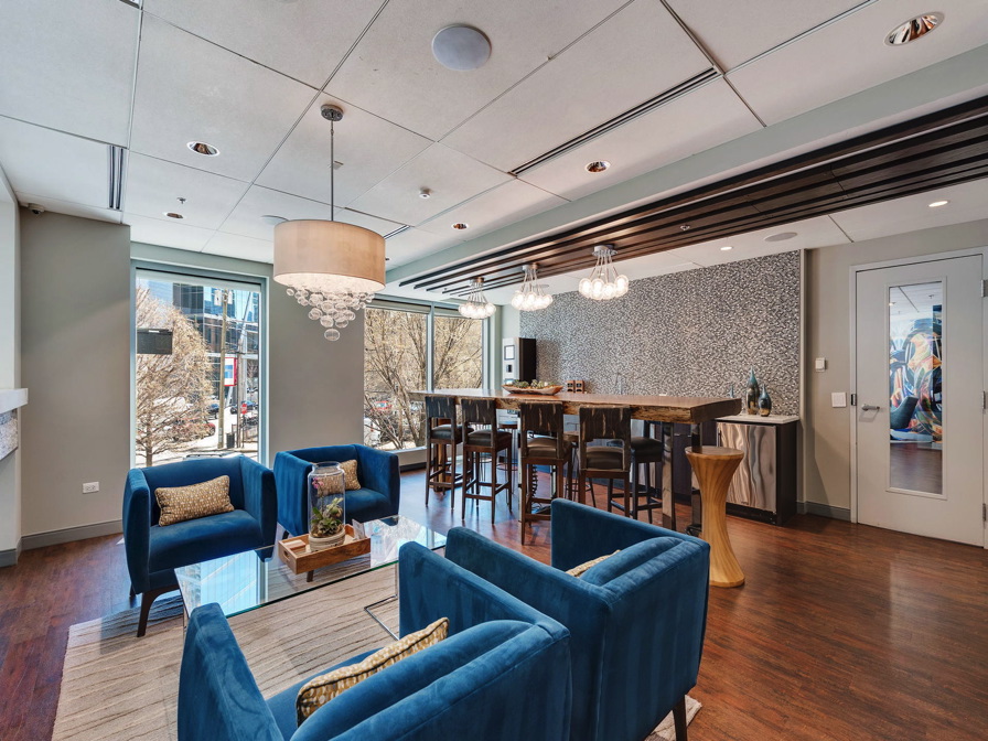 Modern lounge area with blue armchairs, a wooden table, and bar seating. Ceiling lights and large windows add brightness. Contemporary decor with an abstract wall pattern.