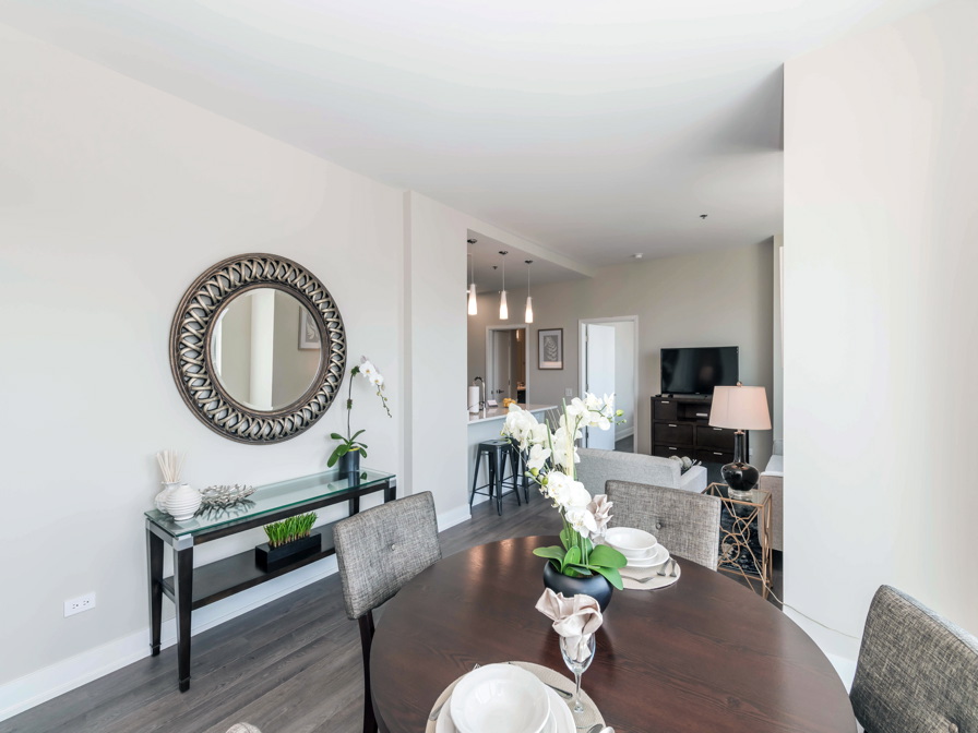 Modern dining and living area with a round table, seating, wall mirror, decorative plants, and a TV in the background. Neutral tones dominate the decor.