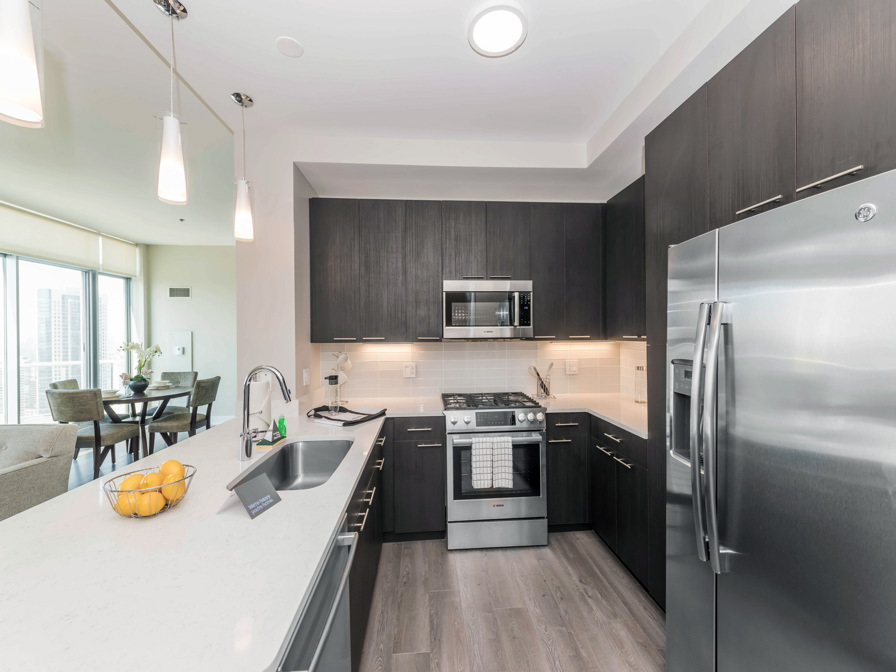 Modern kitchen with stainless steel appliances, dark wood cabinets, and a white countertop. A bowl of lemons sits on the counter. Dining area visible in the background.