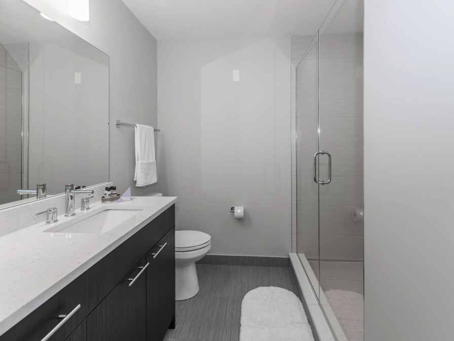 Modern bathroom with glass shower, white vanity with dual sinks, and towel on rack. White bath mat on gray tile floor, under soft overhead lighting.