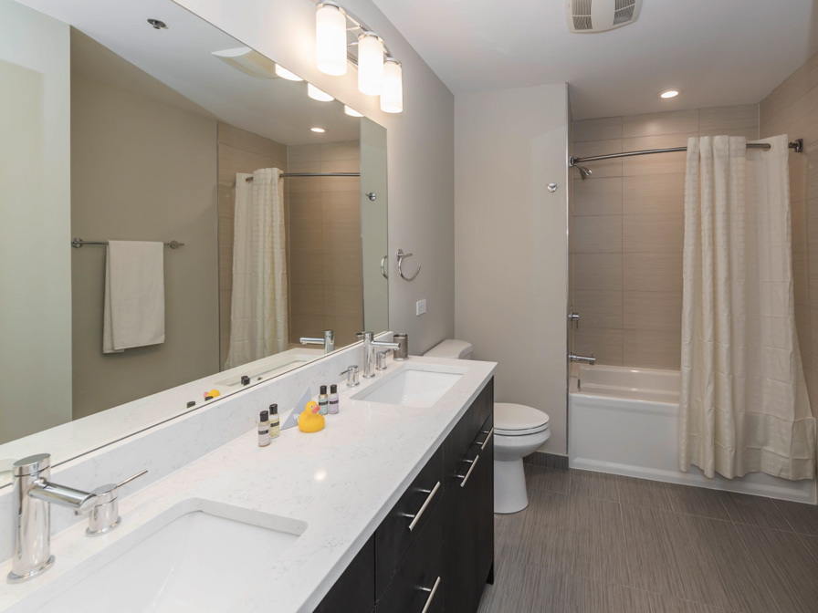 Modern bathroom with double sinks, a large mirror, toiletries, a towel on a rack, a toilet, and a bathtub with a shower curtain.