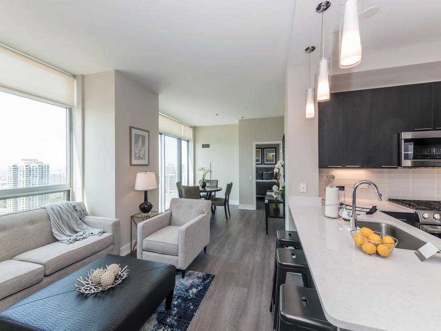 Modern apartment interior with an open layout featuring a kitchen with bar stools, living area with a sofa and armchair, and a distant dining table. Large windows offer a city view.