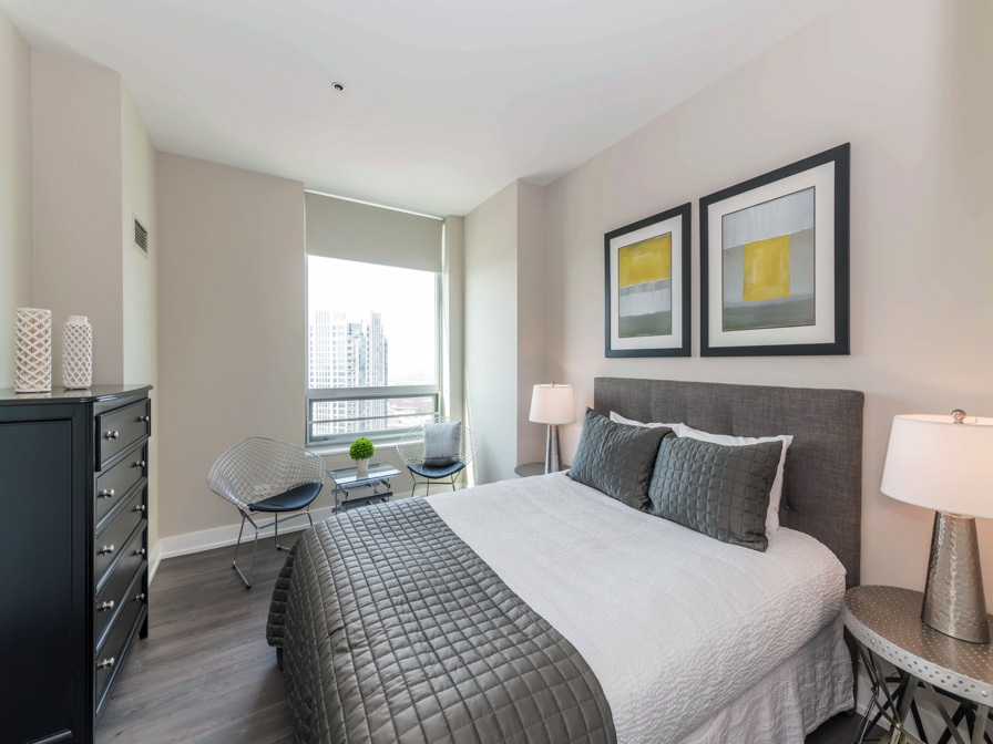 Modern bedroom with a gray and white color scheme, featuring a bed, two framed artworks, a dresser, two chairs by a window, and two lamps. A cityscape is visible through the window.