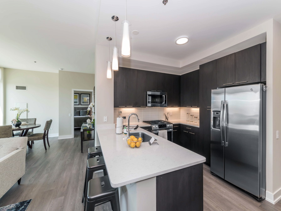 Modern kitchen with dark cabinets, stainless steel appliances, and a white island with hanging lights. Lemons in a bowl sit on the island. Dining area visible in the background.