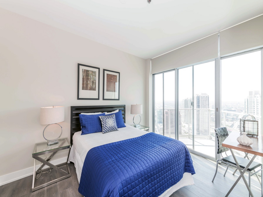 A modern bedroom with a blue and white bed, two side tables with lamps, and large windows overlooking a cityscape. A small table and chair are positioned by the window.